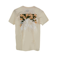Beige t-shirt featuring a detailed back design with an illustration of stairs leading through a garden, overlaid with large orange lilies and surrounded by tropical foliage. A subtle circular logo appears near the top center of the back
