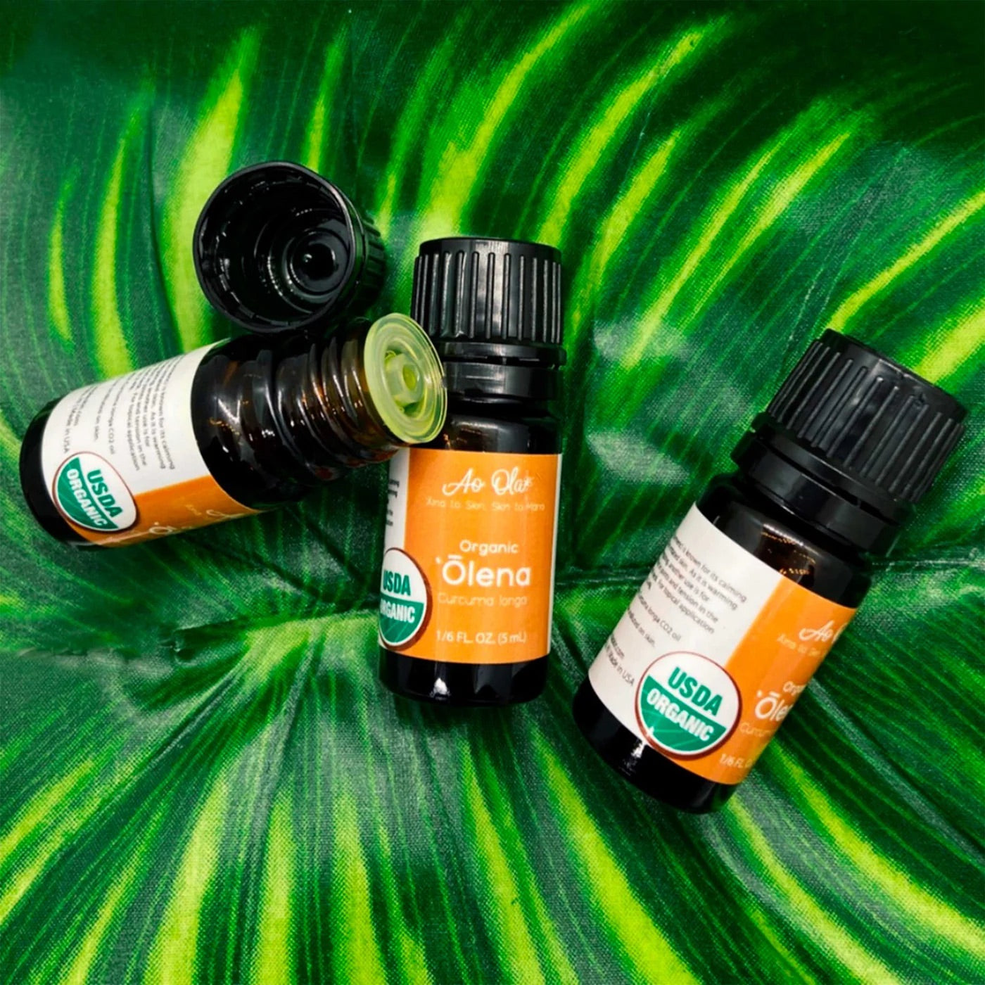 Four ʻōlena essential oil bottles with orange labels against a variegated-leaf background.